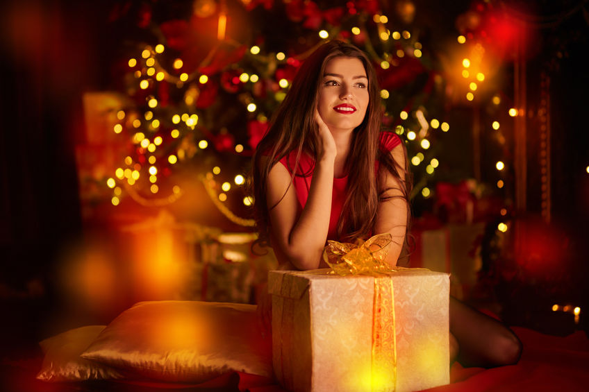 Beautiful dreamy girl sits with a gift box in the fairy Christmas interior with lights around.