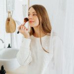 Beautiful smiling young caucasian woman with long hair standing near mirror, holding powder palette and brush, doing makeup and looking at reflection in bathroom at home.