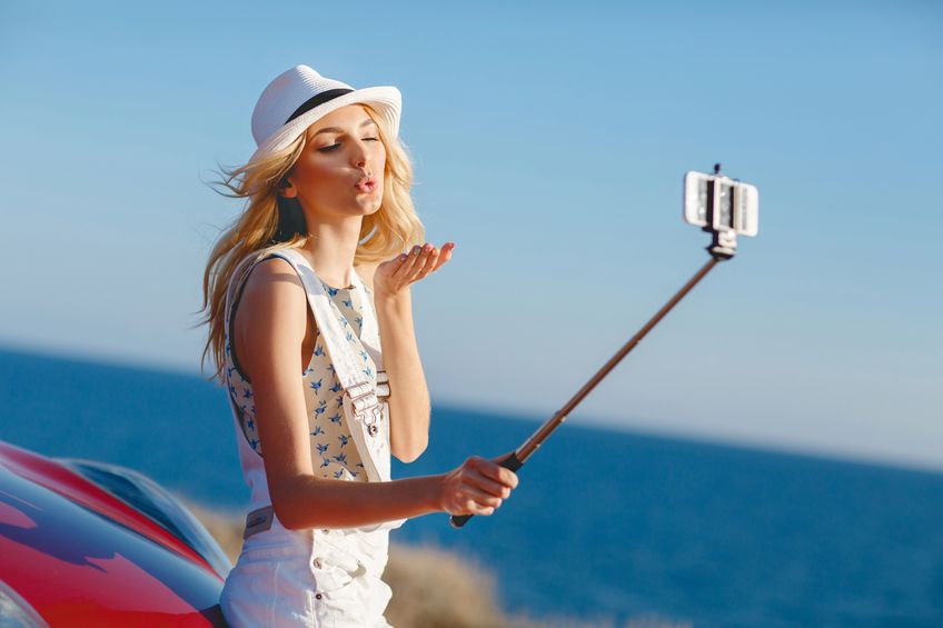 Beautiful, slender woman, with long blonde hair, dressed in a white jumpsuit and a white hat outdoors in summer time, near the red car taking pictures of himself using a smartphone and selfie stick in the background the rocky shore of the ocean